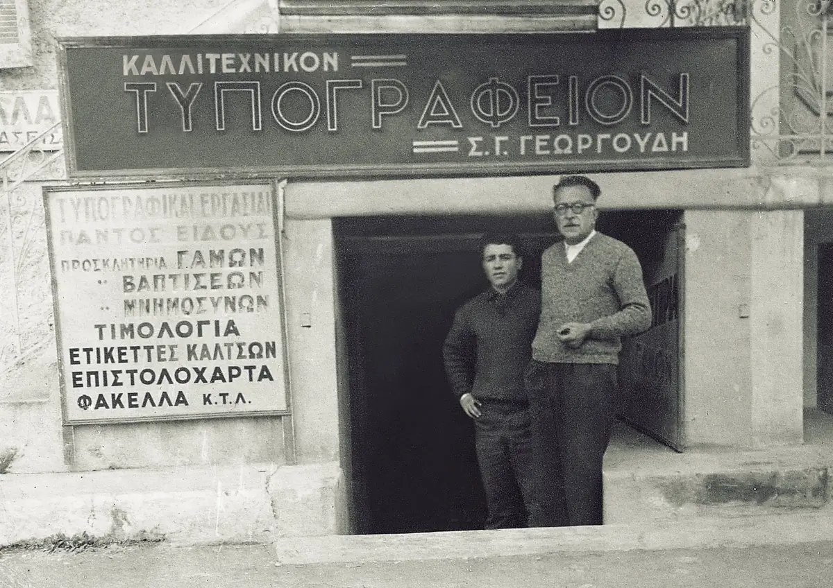 Το τυπογραφείο Σ. Γ. Γεωργούδη: δύο άνδρες στην είσοδο, κάτω από την επιγραφή «Καλλιτεχνικόν Τυπογραφείον»